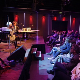 Een volle zaal in Desmet luistert zaterdagavond 15 juni naar een talkshow met host Elma Drayer, die NRC-journalist Marcel Haenen interviewt over zijn boek De Bokser, voorafgaand aan de uitzending van NOS Met het Oog op Morgen.